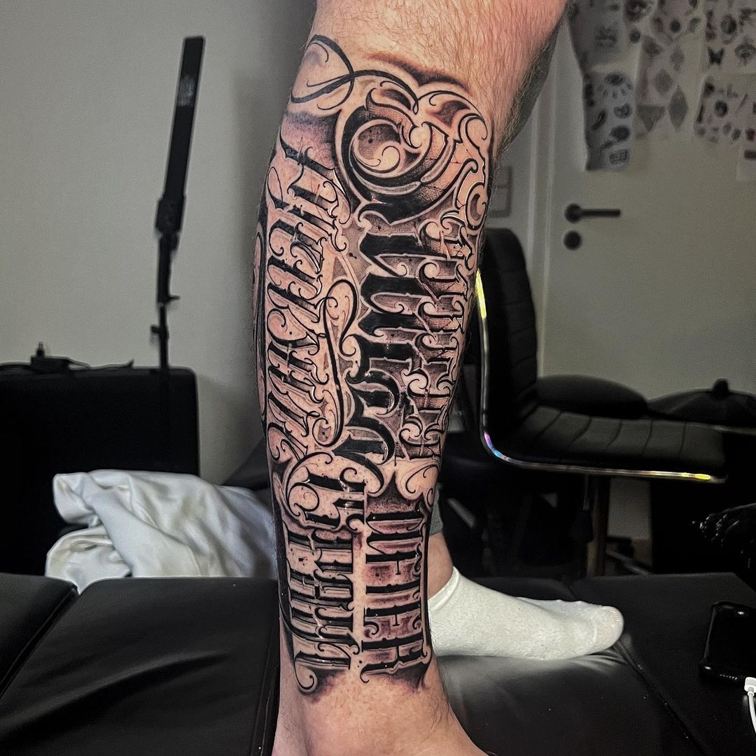 Artistic black and gray tattoo on male leg featuring intricate lettering and swirls.