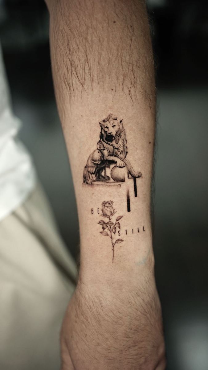 Lion tattoo on forearm with text 'Be Still' and rose illustration
