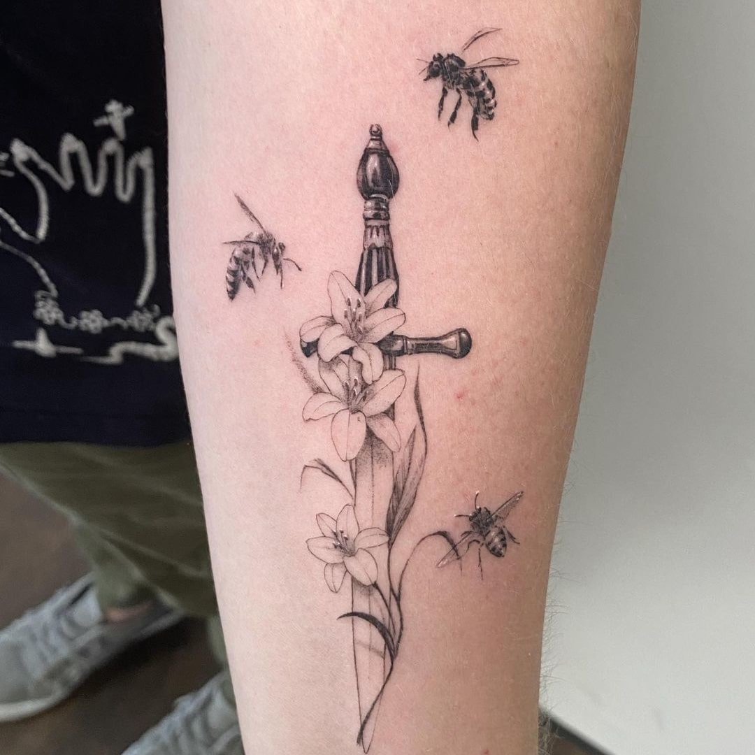 Black and grey tattoo of a sword, flowers, and bees on skin.